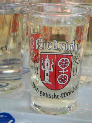 Auf einem Tisch mit einer weiß-blauen Plastiktischdecke  stehen mehrere gefüllte Weinprobiergläser. Auf den Weinprobiergläsern steht in weißer Schrift mit schwarzem Rand "Kiedrich" oberhalb des roten Wappens mit der weißen Burgruine Scharfenstein und dem mit einem Kreuz verbundenem Doppelrad. Unter dem Wappen steht in alter verschnörkelter schwarzer Schrift "das gotische Weindorf".