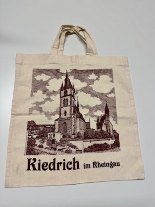 Ein beiger Jutebeutel mit kurzer Schlaufe, auf dem in braun ein viereckiges Bild gedruckt ist, auf dem die Kiedricher Basilica minor, die Kirchhofmauer, die Michaelskapelle, Bänke entlang der Mauer, ein Fachwerkhaus und der Himmel mit Streifen und weißen Wolken dargestellt ist. Darunter sind die Wörter "Kiedrich im Rheingau" in braun gedruckt.
