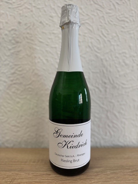 Eine grüne Sektflasche mit weißem Etikett und schwarzer Schrift sowie weißer Alu-Banderole um den Flaschenhals und dem Korken. Die Flasche steht vor einer weiße Strukturtapete.