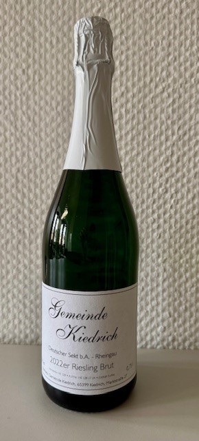Eine grüne Sektflasche mit weißem Etikett und schwarzer Schrift sowie weißer Alu-Banderole um den Flaschenhals und dem Korken. Die Flasche steht vor einer weiße Strukturtapete.