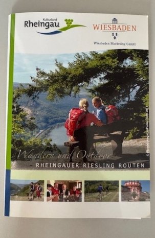 Wanderkarte- auf einer Bank- die unter einem Baum steht, sitzen zwei Wanderer, eine Frau und ein Mann und schauen von weit oben auf das Rheintal. Unter dem großen Bild sind vier weitere kleine Bilder mit einem Fahrrad, eine Menschengruppe, zwei Wanderer und dem Biebricher Schloß in rot und weiß.