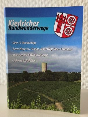Blick auf die Burgruine Scharfenstein, die von grünen Bäumen und Weinbergen umgeben ist. Der Himmel ist blau mit weißen Wolken. Oben rechts ist das Kiedricher Wappen in rot-weiß mit dem Scharfenstein und dem Doppelrad abgebildet.