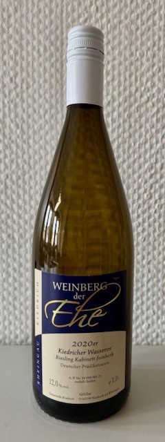 Eine dunkelgrüne Weinflasche, mit beige-blauem Etikett mit weißer, schwarzer und goldener Schrift und weißem Aluschrauber, steht vor einer weißen Strukturtapete.