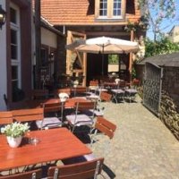 Hof mit Holzstühlen und Tischen mit weißer Blumendeko und weißem aufgespanntem Sonnenschirm vor einem Haus mit großen geöffneten Holztoren.