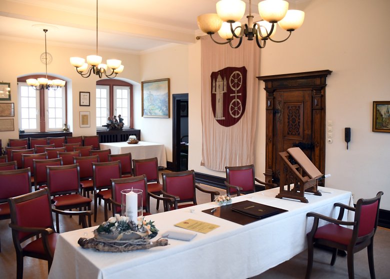 Man sieht das historische Trauzimmer. Rechts ist eine alte und eine modernere Tür und ein Wandteppich in beige mit dem Gemeindewappen in rot-weiß, der Burg Scharfenstein und dem Doppelrad sowie zwei Gemälde mit goldenem Rahmen. An der anderen Wand sieht man zwei Sprossenfenster und drei Kronleuchter mit je 6 Lampenschirmen. Die Stühle sind aus braunem Holz und die Sitz- und Rückenpolster aus weinrotem Leder. Vorne ist ein langer Trautisch mit beiger Tischdecke, einer beigen Kerze, ein ovales Blumengesteck, einer beigen Urkunde, einem Kugelschreiber, ein Blumenherz für die Ringe und ein Pult aus braunem Holz mit einer hellbraunen Mappe.