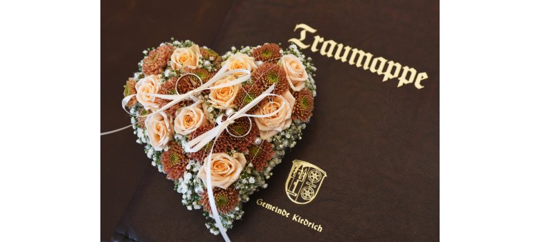 Auf einer braunen Ledermappe ist mit goldener Schrift "Traumappe" und "Gemeinde Kiedrich" sowie das Wappen mit der Burg Scharfenstein und dem Doppelrad abgebildet. Darauf liegt ein Herz, aus lachsfarbenen Rosen und braunen Chrysanthemen, akzentuiert mit Schleierkraut und weißen Perlen sowie silbernem gezacktem Draht. Auf dem Herz liegen zwei Ringe, die jeweils mit einem beigen Band , das zu einer Schleife gebunden ist, befesigt sind.