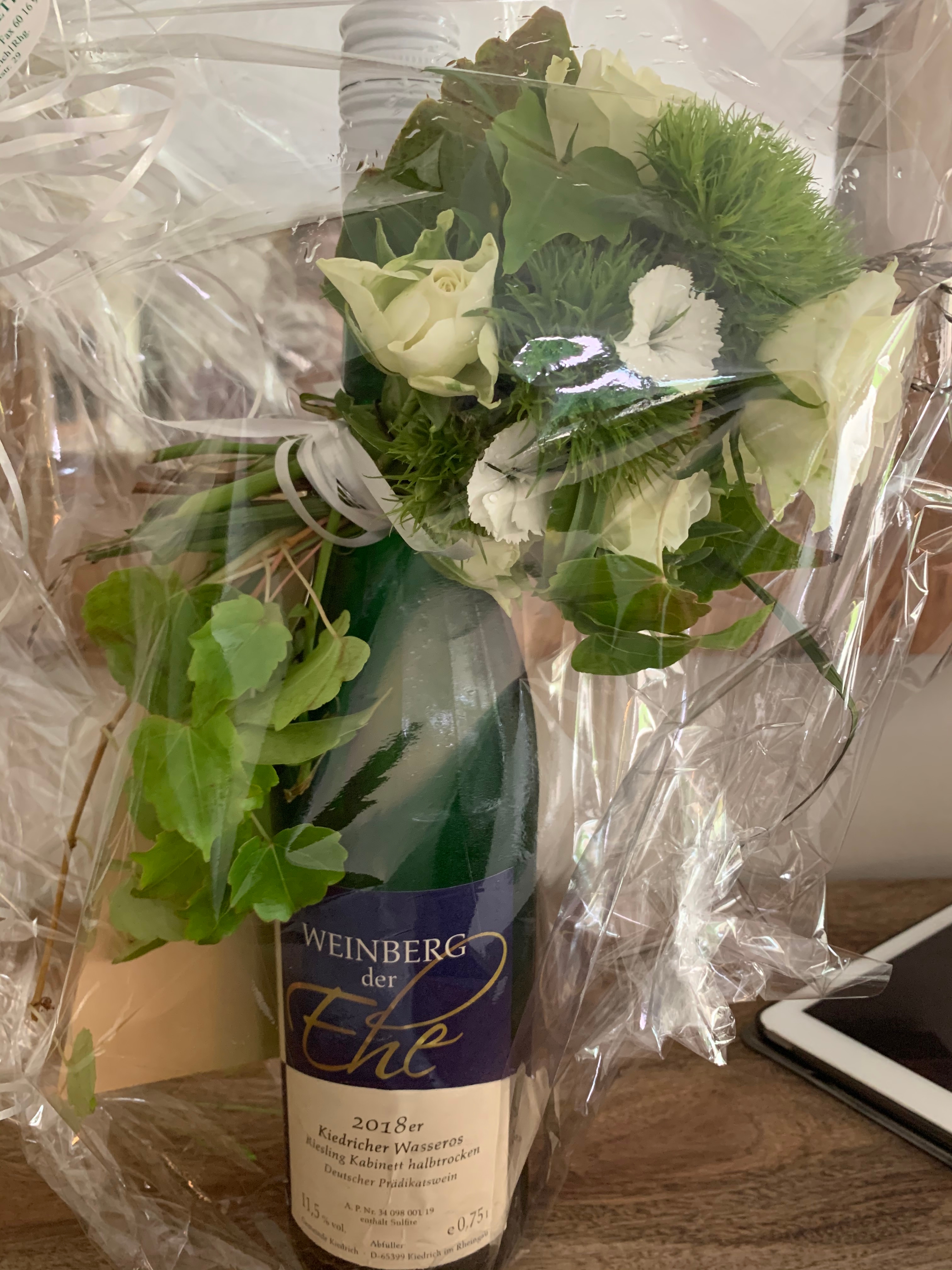 Weinflasche aus dem "Weinberg der Ehe" Eine in Klarsichtfolie eingepackte grüne Flasche Wein aus dem Weinberg der Ehe mit dem blau-beiges Weinetikett Weinberg der Ehe. Oben in blau mit silberner geschwungener Schrift "Ehe" und unterhalb in dunkler Schrift auf beigem Hintergrund der Jahrgang, die Menge, die Inhaltsstoffe und die Weinlage beschrieben. Am Flaschenhals wurde ein kleiner Blumenstraße mit weißen Rosen und weißen Blumen und grünen Gräsern und Blättern gebunden.