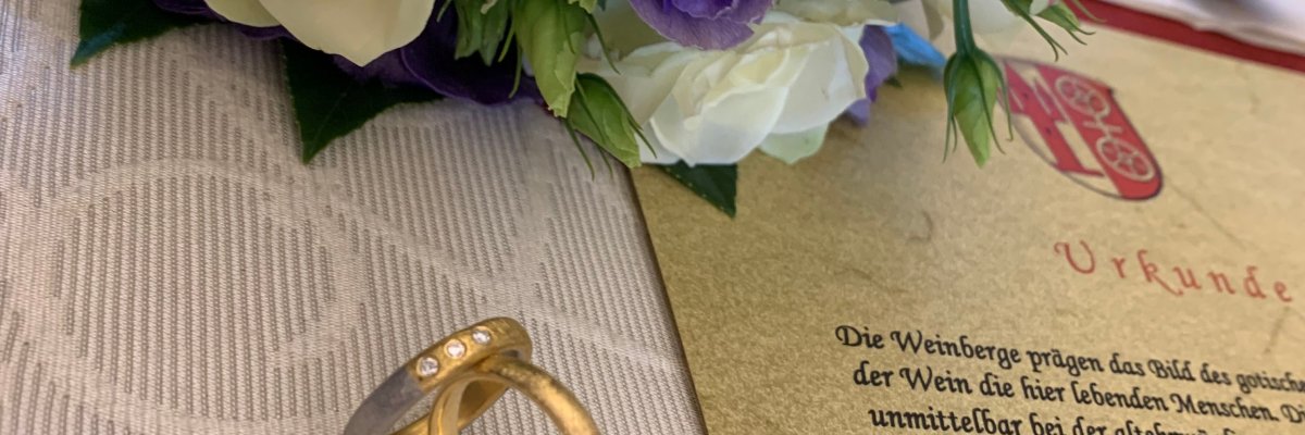 Rechts liegt die auf elefantenbogen-Papier gedruckte Urkunde Weinberg der Ehe. Links davon sind die aufgestellten Eheringe in Gold, einer davon mit drei Steinen verziert. Sie befinden sich auf einer weißen Stofftischdecke mit Schnörkeln. Oberhalb liegt der Brautstrauß mit vier weißen Rosen und lila Blumen. 
