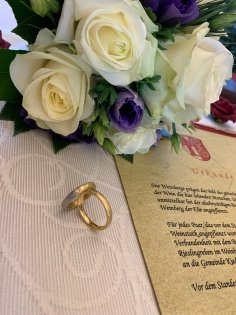 Rechts liegt die auf elefantenbogen-Papier gedruckte Urkunde Weinberg der Ehe. Links davon sind die aufgestellten Eheringe in Gold, einer davon mit drei Steinen verziert. Sie befinden sich auf einer weißen Stofftischdecke mit Schnörkeln. Oberhalb liegt der Brautstrauß mit vier weißen Rosen und lila Blumen. 