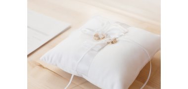 Auf einer hellen Holzunterlage liegt ein weißes Ringkissen mit weißen Bändern, mit dnen zwei goldene Eheringe befestigt sind. Links davon liegt ein weißes Blatt.