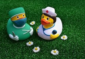 Auf einem Kunstrasen liegen die Blüten von fünf Gänseblümchen und zwei Badeenten. Eine weibliche Ente hat  einen weißen Kittel, eine weiße Haube mit rotem Kreuz und ein Stethoskop um den Hals und in der Hand. Die andere Ente hat einen grünen Kittel, eine grüne OP-Haube,  Handschuhe, eine Mundschutz an und ein Messer in der einen und eine Schere in der anderen Hand. 