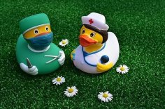 Auf einem Kunstrasen liegen die Blüten von fünf Gänseblümchen und zwei Badeenten. Eine weibliche Ente hat  einen weißen Kittel, eine weiße Haube mit rotem Kreuz und ein Stethoskop um den Hals und in der Hand. Die andere Ente hat einen grünen Kittel, eine grüne OP-Haube,  Handschuhe, eine Mundschutz an und ein Messer in der einen und eine Schere in der anderen Hand. 