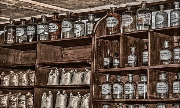 Apotheke Blick in ein altes braunes Holzregal, welches mit alten Apotheker-Glasflaschen mit jeweils weißen ovalen Flaschenetiketten mit Aufdruck bestückt ist. Ebenso stehen silberne beschriftete Säckchen in den Regalen.