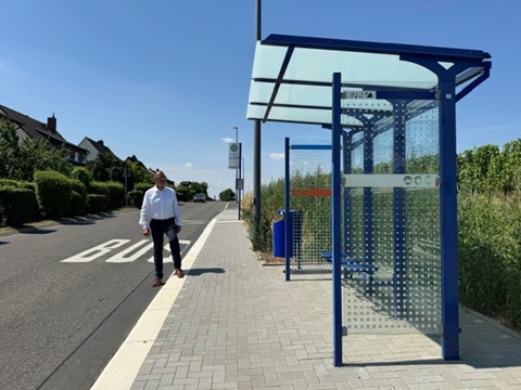 Vor Weinbergen steht eine blaue Bushaltestelle mit Glaswänden und Glasdach. Die Bordsteine sind erhöht und auf der Straße steht "BUS" und am Bürgersteig ist ein Schild in weiß mit einem  grünem H und drum herum ein grünem Kreis mit gelber Füllung. Auf dem Bürgersteig steht ein Mann in dunkler Hose und weißem Hemd.