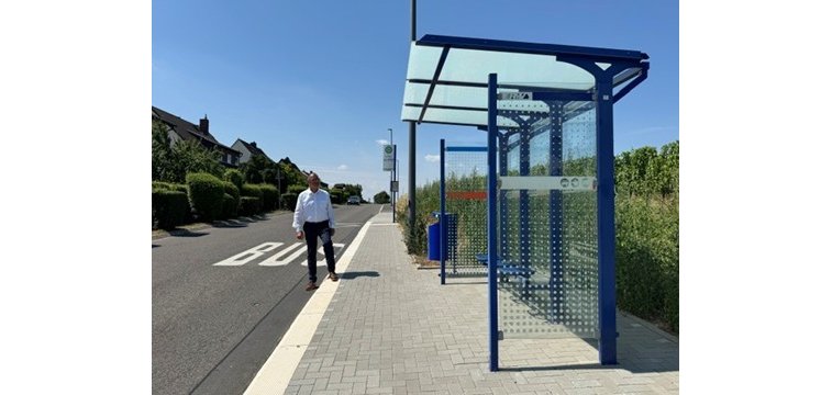 Vor Weinbergen steht eine blaue Bushaltestelle mit Glaswänden und Glasdach. Die Bordsteine sind erhöht und auf der Straße steht "BUS" und am Bürgersteig ist ein Schild in weiß mit einem grünem H und drum herum ein grünem Kreis mit gelber Füllung. Auf dem Bürgersteig steht ein Mann in dunkler Hose und weißem Hemd.