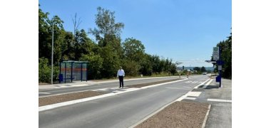 Auf einer breiten Straße mit zwei Fahrbahnen ist in der Mitte eine neue Verkehrsinsel mit Erde, die noch zu bepflanzen ist. Dort steht ein Mann in dunkler Hose und weißem Hemd. Links und rechts der Fahrbahn sind neue blaue Bushaltestellen mit Glaswänden nd barrierefreien Bürgersteigen.