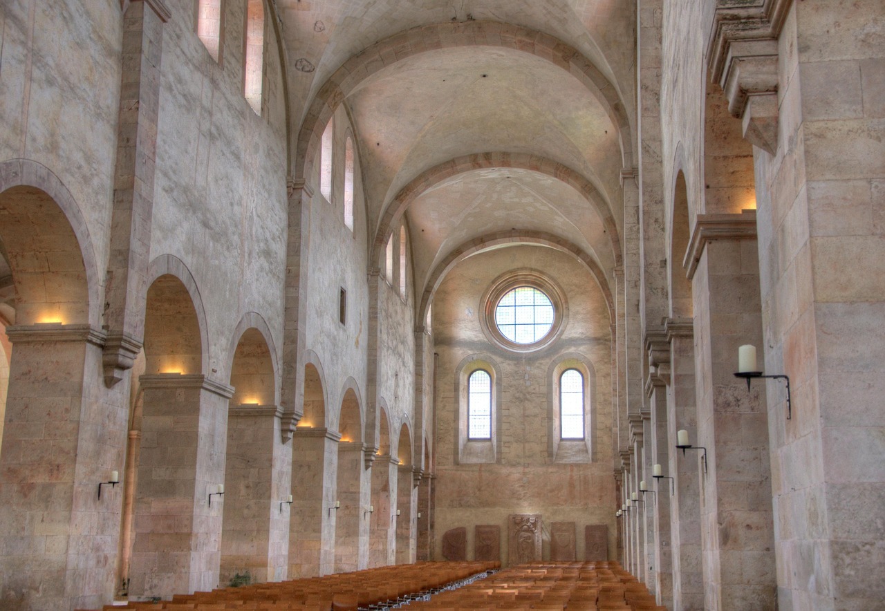 Ein Kirchenschiff, mit hohen grauen Mauern und Bögen an der Decke. Links und rechts sind breite Säulen mit hohen Bögen und jweweils ein schwarzer Kerzenständer mit beigen Kerzen drauf. In den Säulen scheint ein Licht in dem Bereich, an dem die Säulen beginnen. Hinten sind zwei Fenster, die oben ebenso einen Bogen haben und darüber ein kreisförmiges Fenster. Unterhalb der Fenster sind fünf rötliche, stehende Grabplatten. Die Basilika ist mit braunen Holzstühlen und einem Mittelgang bestuhlt.