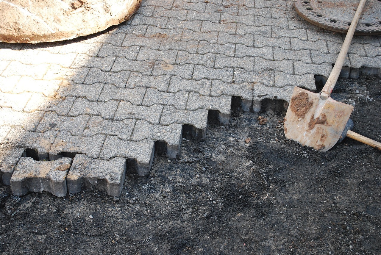 Ein Geweg mit Pflastersteinen, auf denen ein runter Gullideckel liegt. Vorn sind die Steine entfernt und man sieht Split und zwei Schaufeln. 