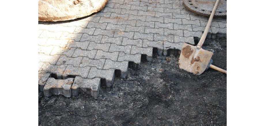 Ein Geweg mit Pflastersteinen, auf denen ein runter Gullideckel liegt. Vorn sind die Steine entfernt und man sieht Split und zwei Schaufeln. 