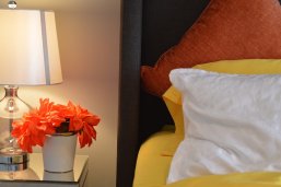Auf einem Bett , welches mit einem gelben Spannbettlaken bezogen ist, liegt ein weißes, ein gelbes und ein orangenes Kissen. Neben dem Bett steht ein weißer Nachttisch mit einer Glasplatte, auf dem orangene Blumen in einem weißen Blumentopf mit einem silbernen Rand stehen. Daneben steht eine Tischlampe mit einem weißen Lampenschirm.