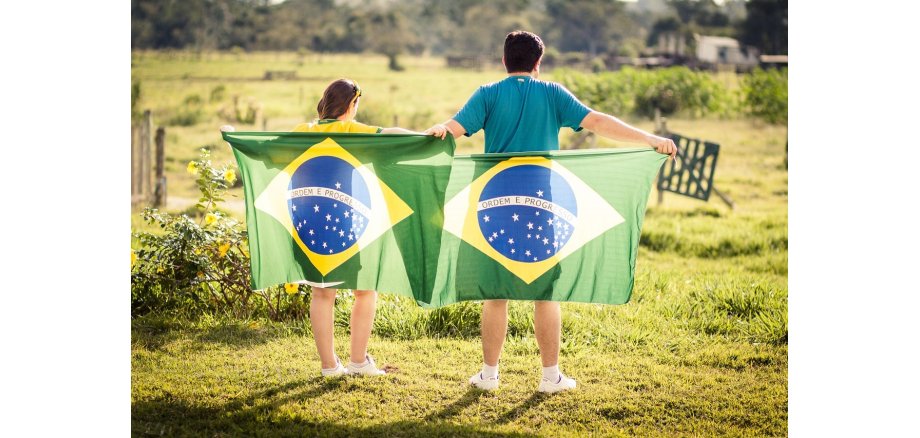 Auf einer Wiese mit verschwommenen Gattern und Bäumen im Hintergrund steht ein jugendlicher Junge und ein Mädchen mit kurzen Hosen, mit dem Rücken zum Fotografen. Beide halten je eine Brasilienfahne mit ausgestreckten Armen hinter ihren Rücken. Die Fahne ist grün mit einer gelben Raute, in der eine blaue Kugel mit gelben Sternen und einem Schriftzug ist.