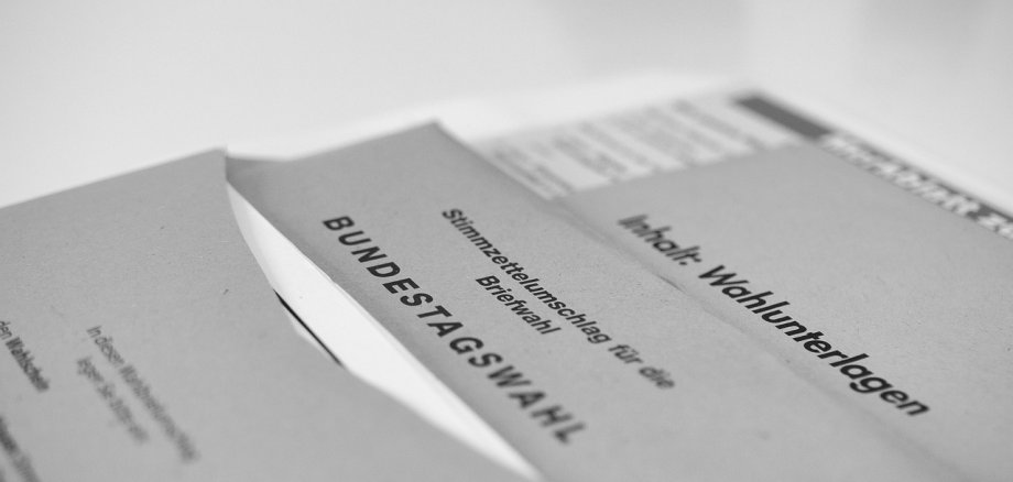 Auf einem grauen Tisch liegen mehrere graue Briefumschläge mit der Beschriftung Bundestagswahl und einem weißem Zettel. Ganz unten im Stapel ist ein Formular.