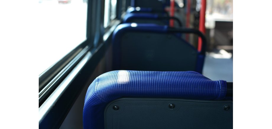 Man sieht verschwommen einen Ausschnitt des Inneren eines Linienbusses, d.h. vier Sitze mit blauem Stoff und grauer Rückseite und rote Haltestangen. Links ist eine breite Fensterreihe zu erkennen.