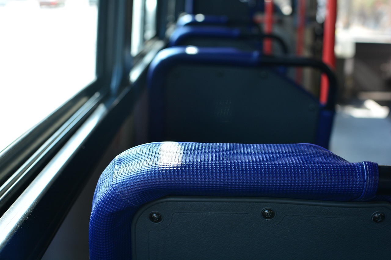Man sieht verschwommen einen Ausschnitt des Inneren eines Linienbusses, d.h. vier Sitze mit blauem Stoff und grauer Rückseite und rote Haltestangen. Links ist eine breite Fensterreihe zu erkennen.
