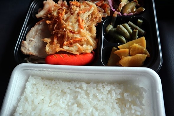 Eine rechteckige weiße Styroporschale, die mit weißem Reis gefüllt ist. Oben drüber ist eine schwarze rechteckige Plastikschale, die mit chinesischem Essen, Fleisch, Gemüse und einer weiteren schwarzen, rechteckigen, kleineren Plastikschale gefüllt ist.   Die Schalen stehen auf einem Schwarzen Untergrund.