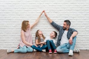 Vor einer weißen gemauerten Wand, auf einem braunen Holzboden, sitzt eine Familie. Links die Mutter, daneben ein Mädchen, danaben ein Junge und dann der Vater. Die Mutter und der Vater geben sich die Hand und formen sie zu einem Dach. Die Kinder schauen nach oben zu der Spitze, die Eltern schauen zu den Kindern.