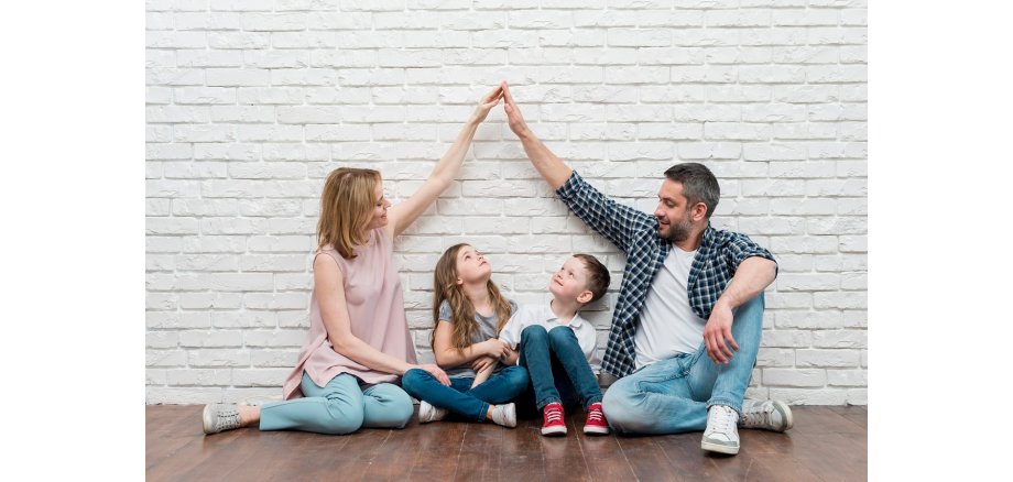 Vor einer weißen gemauerten Wand, auf einem braunen Holzboden, sitzt eine Familie. Links die Mutter, daneben ein Mädchen, danaben ein Junge und dann der Vater. Die Mutter und der Vater geben sich die Hand und formen sie zu einem Dach. Die Kinder schauen nach oben zu der Spitze, die Eltern schauen zu den Kindern.