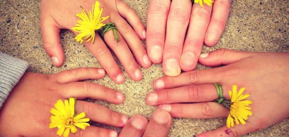 Fünf Hände, von Erwachsenen und Kindern, liegen auf einem Untergrund aus Sand. Die Fingerspitzen zeigen zueinander und die Hände bilden einen Kreis. An den Mittelfingern ist ein gelb blühender Löwenzahn und der grüne Stiel der Blume ist um den Mittelfinger gewickelt.
