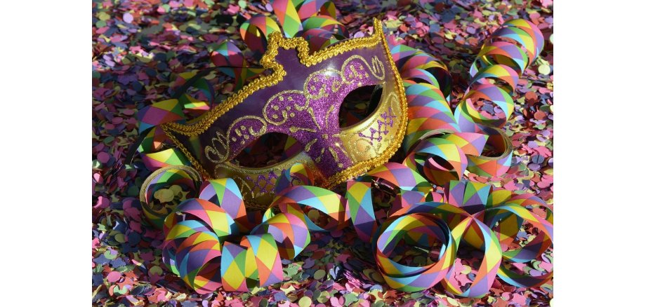 Eine Augenmaske, mit einer goldenen Bordüre und lila Glitzer mit goldenene Verzierungen über den Augen und goldenem Glitzer mit lila- und goldenem Glitzer-Verzierungen unter den Augen, liegt auf buntem Konfetti, auf dem auch Luftschlangen liegen, die Rauten in lila, hellblau, gelb, orange und grün zeigen.  