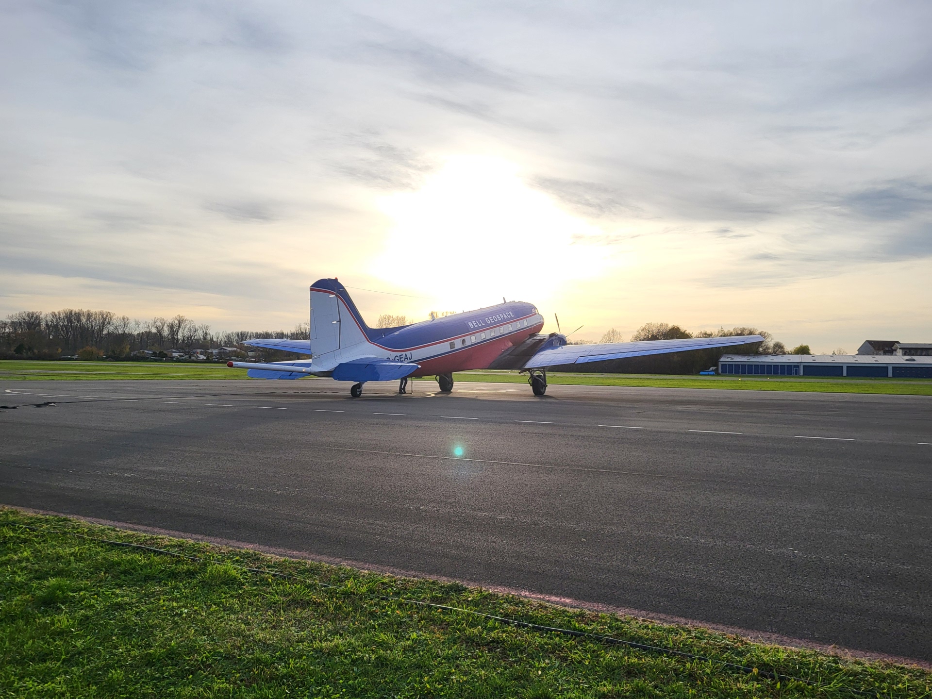 Auf einer asphaltierten Landebahn, die von Rasen umgeben ist, steht ein Kleinflugzeug in blau, rot, weiß. Im Hintergrund ist ein Tanga und der Himmel ist bewölkt. 