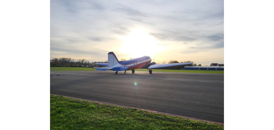 Auf einer asphaltierten Landebahn, die von Rasen umgeben ist, steht ein Kleinflugzeug in blau, rot, weiß. Im Hintergrund ist ein Tanga und der Himmel ist bewölkt.