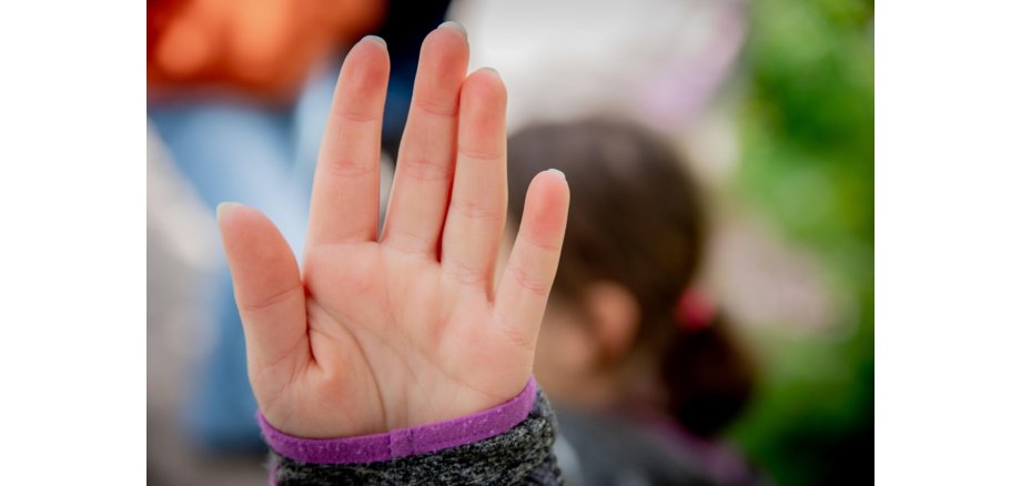Im Vordergrund eine Hand, die nach vorn ausgestreckt ist. Man sieht die Handinnenfläche und ein Stück von einem grau melierten Pulli mit lila Rand. Im Hintergrund, verschwommen eine Frau mit schulterlagen, dunklen Haaren, die zu einem Pferdeschwanz gebunden sind.