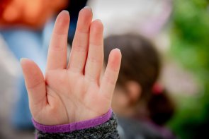 Im Vordergrund eine Hand, die nach vorn ausgestreckt ist. Man sieht die Handinnenfläche und ein Stück von einem grau melierten Pulli mit lila Rand. Im Hintergrund, verschwommen eine Frau mit schulterlagen, dunklen Haaren, die zu einem Pferdeschwanz gebunden sind.
