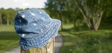 Auf einem Holzpfosten hängt eine Kindermütze aus Jeansstoff mit weißen Erdbeerblüten und roten Erdbeerfrüchten. Im Hintergrund ist verschwommen ein grauer Weg zu erkennen, der durch eine gemähte Wiese läuft. Ebenso sind im Hintergrund Laubbäume verschwommen dargestellt.