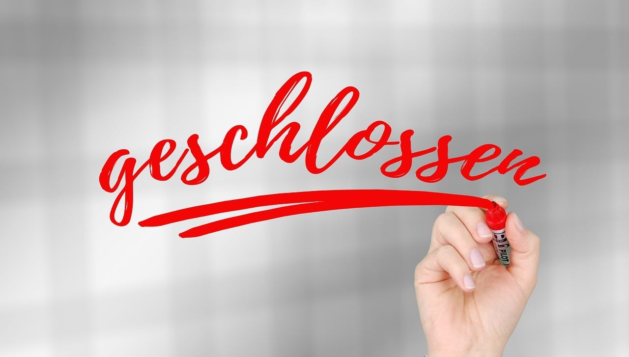 Eine Hand schreibt mit einem roten Edding das Wort "geschlossen" und unterstreicht es doppelt. Der Hintergrund ist verschwommen, weiß, grau, karriert.