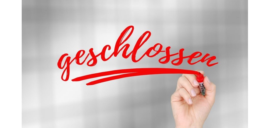 Eine Hand schreibt mit einem roten Edding das Wort "geschlossen" und unterstreicht es doppelt. Der Hintergrund ist verschwommen, weiß, grau, karriert.