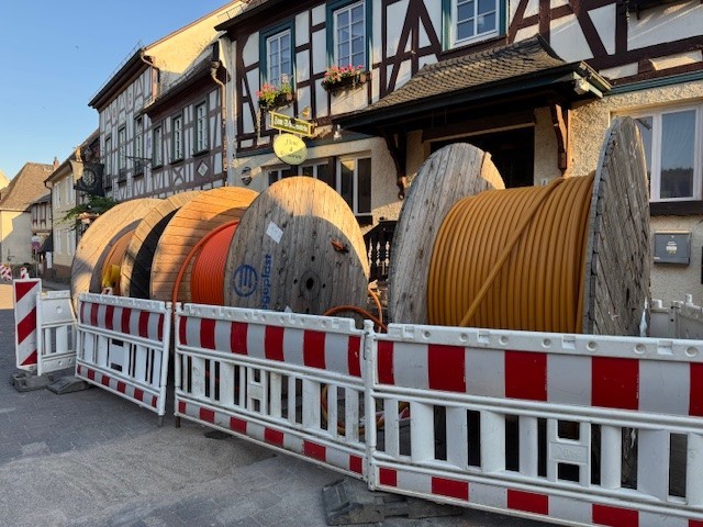 Vor mehreren Fachwerkhäusern stehen drei große Spuolen aus Holz, auf denen Glasfaserkabel in gelb und orange aufgerollt ist. Davor sind vier Warnbaren in rot-weiß.