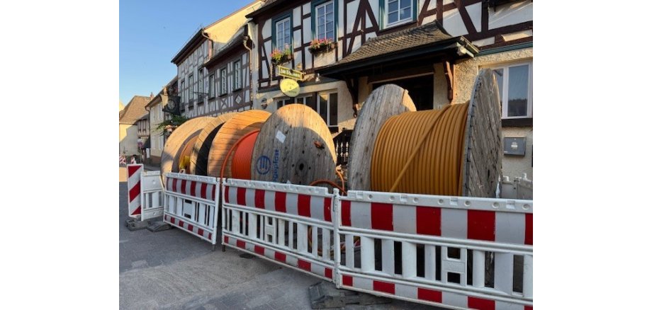 Vor mehreren Fachwerkhäusern stehen drei große Spuolen aus Holz, auf denen Glasfaserkabel in gelb und orange aufgerollt ist. Davor sind vier Warnbaren in rot-weiß.
