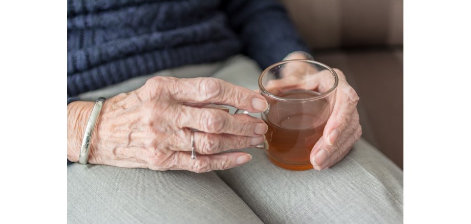 Man sieht die Hände einer älteren Frau, die ein Glas mit Tee hält, welches auf dem Bein steht. Sie trägt eine graue Hose und einen blauen Strickpullover.