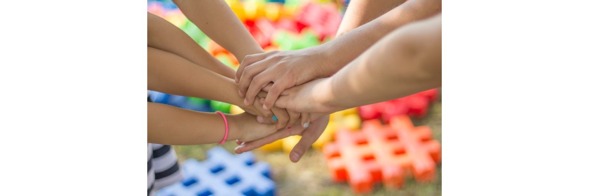 Sieben Hände stapeln sich übereinander. Es sind ebenso die nackten Arme zu sehen. An einem Arm ist ein pinker Haargummi und die Nägel einer Hand sind pink, weiß und hellblau lackiert. Im Hintergrund sind auf einem Boden verteilte Raster mit je drei Balken, die jeweils einfarbig sind. Es gibt Orangene, Gelbe, Grüne, Blaue und Rote.