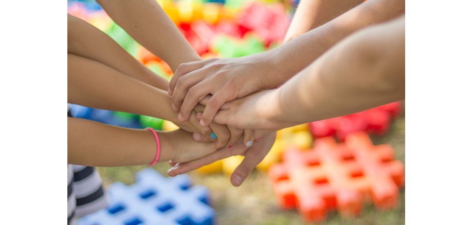 Sieben Hände stapeln sich übereinander. Es sind ebenso die nackten Arme zu sehen. An einem Arm ist ein pinker Haargummi und die Nägel einer Hand sind pink, weiß und hellblau lackiert. Im Hintergrund sind auf einem Boden verteilte Raster mit je drei Balken, die jeweils einfarbig sind. Es gibt Orangene, Gelbe, Grüne, Blaue und Rote. 
