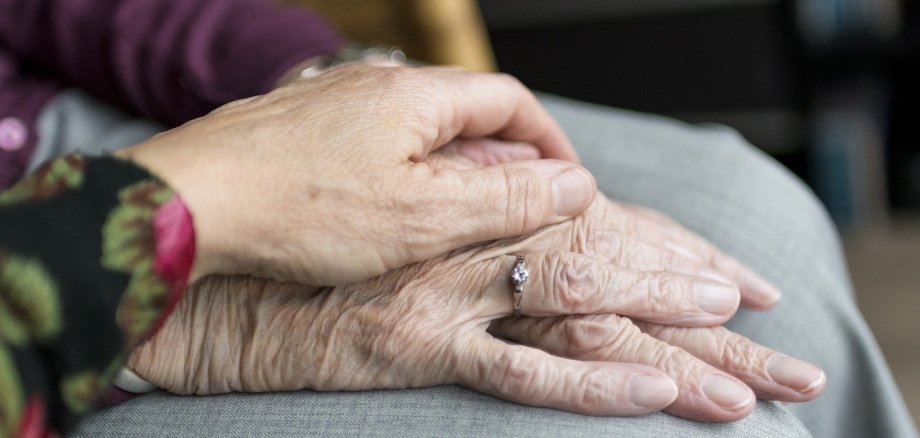Eine Frau sitzt auf einem hellbraunen Stuhl. Sie hat eine hellgraue Hose und einen lila Pullover an. Man sieht keinen Körper und kein Gesicht.  Ihre Hände, die Falten haben, liegen aufeinander auf ihren Beinen. An dem Ringfinger steckt ein silberner Ring mit einem Stein. Eine weitere Hand, ebenfalls mit Falten, liegt augf den Händen der anderen Frau. Der Ärmel ist schwarz mit pink-roten runden Blüten, die mit runden grün-braunen Blättern umrandet sind.