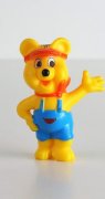 Eine gelbe Gummibärenfigur steht auf einer weißen Fläche. Er hat eine blaue Latzhose an und ein rotes Halstuch und ein rotes Band um die Stirn. 