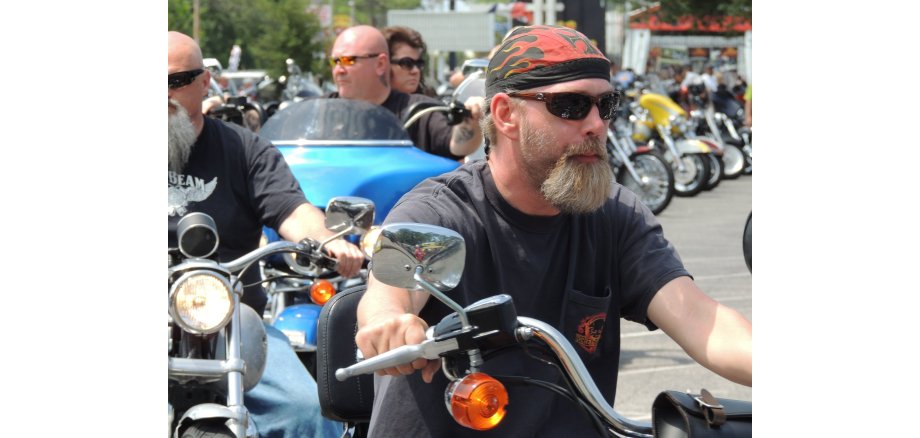 Man sieht drei Männer und eine Frau als Mitfahrerin auf drei Motorrädern mit Sonnenbrillen. Zwei der Männer haben lange Bärte, einer ein Flammenkopftuch und zwei Glatzen. Im Hintergrund sind weitere Motorräder. 