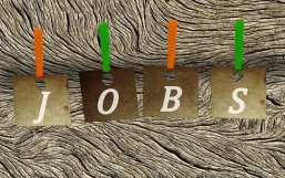 Jobs Im Hintergrund ist ein Stück Holz. Davor sind vier braune viereckige Zettel mit Loch oben. Auf jedem Zettel steht ein weißer Großbuchstabe "JOBS". Die Zettel haben oben jeweils eine Klapper, insgesamt zwei orangene und zwei grüne Klappern.