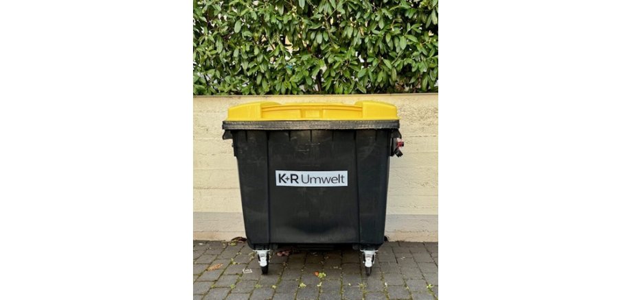 Vor einer Mauer, die unten einen grauen Streifen hat und darüber beige ist, stehet eine große Mülltonne. Sie ist schwarz und hat einen gelben rechteckigen Deckel. Auf der Tonne ist ein weißes Rechteck, in dem in schwarz "K+R Umwelt" steht. Über der Mauer ist eine Hecke mit grünen ovalen Blättern. 